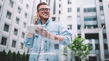 Kot ceketli, kulaklıklı, modern tablet kullanan ve sokakta mutlu bir şekilde gülümseyen mutlu bir adamdan.