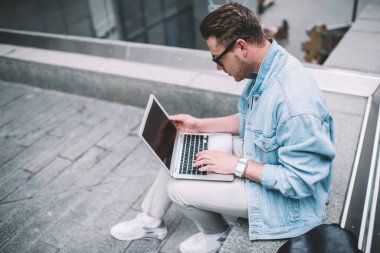 Gözlüklü ve kot ceketli havalı beyaz adam sokak çitlerinde oturuyor ve taşınabilir laptop üzerinde çalışıyor.