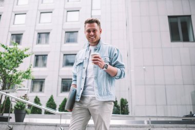 Sokağın ortasında kameraya gülümseyen, tek kullanımlık kahve ve dizüstü bilgisayarlı yakışıklı genç adamdan.