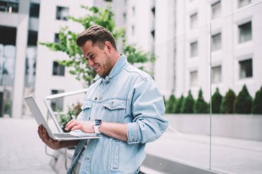 Çağdaş şehir sokağı arka planında dururken, kot ceketli gülümseyen bir adamın taşınabilir dizüstü bilgisayarda daktilo edişinin yan görüntüsü