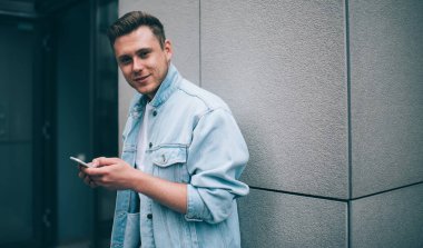 Şık kot ceketli, genç ve yakışıklı bir adam duvara yaslanmış ve kameraya bakarken cep telefonunu tutuyor.