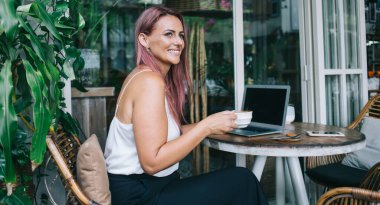 Yetişkin bir kadının kafede oturmuş, kahve içerken bilgisayarlı akıllı telefonu iş için kullanırken ve başka tarafa bakarken yan görüntüsü.