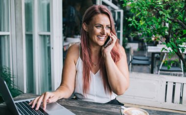 Yazın terasta kahve fincanı açık otururken akıllı telefondan konuşan ve dizüstü bilgisayarda yazan mutlu bir kadın.