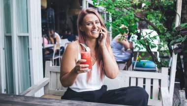 Elinde smoothie ile kafedeki beyaz ahşap bankta oturan telefonda kameraya bakarak konuşan güzel, neşeli bir kadın.