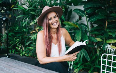 Çekici, şapkalı, saçları boyalı, kitap okuyan, tropik çalıların yanındaki masada oturan ve kameraya bakan neşeli bir kadın.