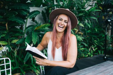 Çekici, şapkalı, boyalı saçlı, kitap okuyan, tropik çalıların yanında oturan ve kameraya bakan bir kadın.