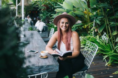 Kafe terasında kitap okurken elinde kahve fincanıyla oturmuş kameraya bakan şapkalı çekici bir kadın.