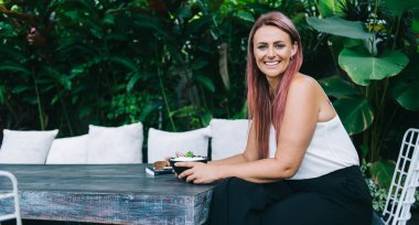 Tarz sahibi pembe saçlı, neşeli yetişkin bir kadın. Dostça gülümseyen ve kameraya bakan. Tropikal bahçede kahvaltıda sağlıklı yulaf lapasının tadını çıkaran.