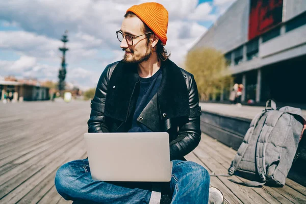 Portrait of trendy dressed hipster guy sitting with laptop computer for e learning outdoors satisfied with 4G internet connection, prosperous male freelancer enjoying sunny day on publicity area