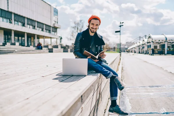 Portrait of cheerful caucasian male student spending time for preparing to university examination sitting in campus with laptop computer and textbook for education, positive hipster guy with notepad