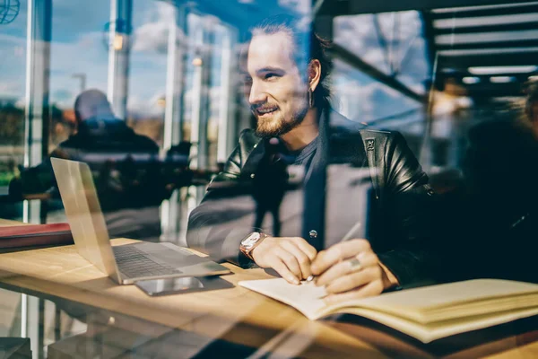 正のCaucasian男に座っている窓の近くのカフェでラップトップコンピュータと教科書作業オンライン 明るい笑顔男性ブロガー支出時間でカフェテリアでリモート仕事をする上でネットブック経由で — ストック写真
