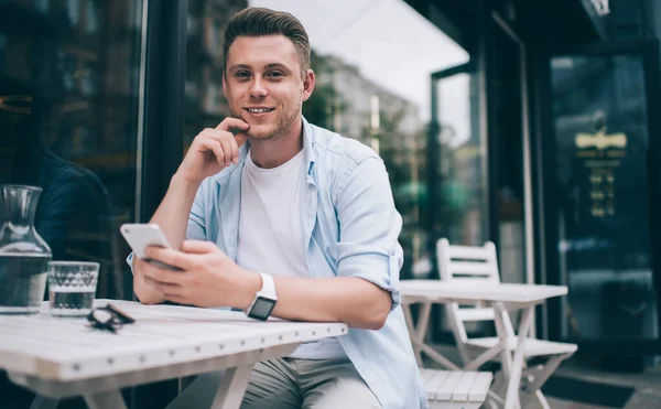 Contemporary millennial man in casual clothes sitting at white table outside holding smartphone and smiling happily at camera on street