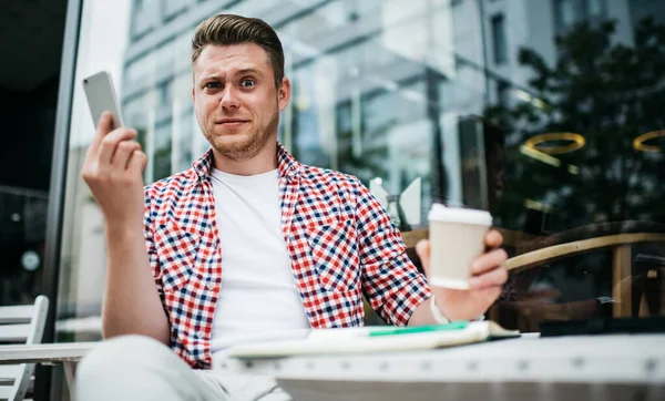 From below of millennial grimacing man holding phone and coffee cup while looking at camera with expression of confusion sitting on street