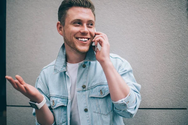 Handsome trendy man in denim jacket and smart watch standing on street and smiling happily while having phone call gesturing happily
