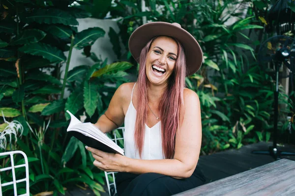 Çekici, şapkalı, boyalı saçlı, kitap okuyan, tropik çalıların yanında oturan ve kameraya bakan bir kadın.
