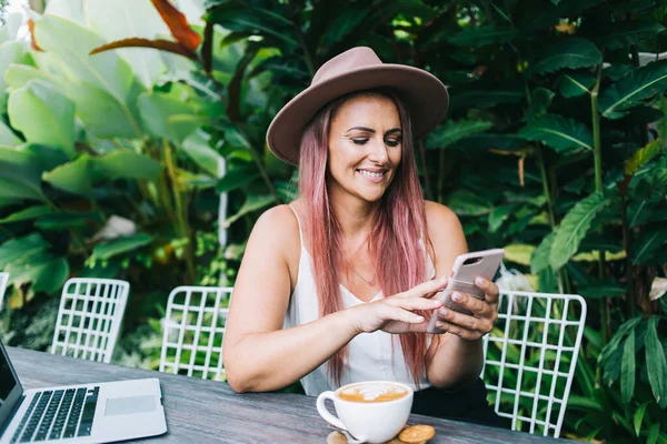 Smiling freelancer with hat on head sitting in garden with cup of aromatic coffee on table and browsing social media during break