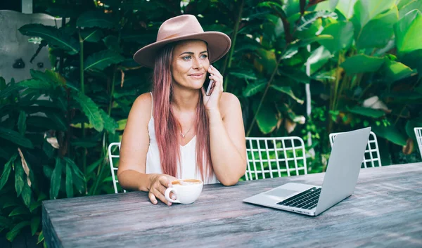 Pozitivní Usmívající Žena Klobouku Mluví Smartphone Prohlížení Notebooku Zatímco Sedí — Stock fotografie
