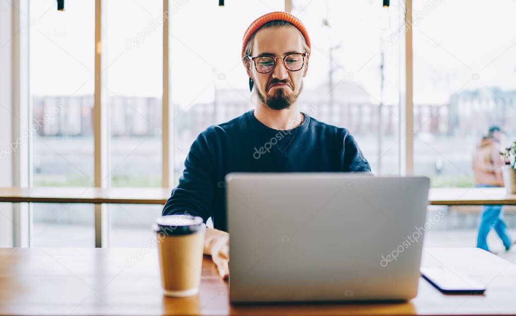 Male freelancer feeling envy while checking rating of successful workers of month on website of company connected to public wifi internet on laptop device, hipster guy watching online video