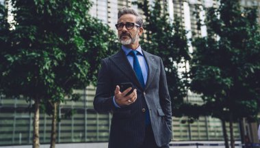 Orta yaşlı, gri saçlı, takım elbiseli, gözlüklü, modern dijital akıllı telefon kullanan iş adamı ağaçlarla çevrili kentsel arka plana bakıyor.