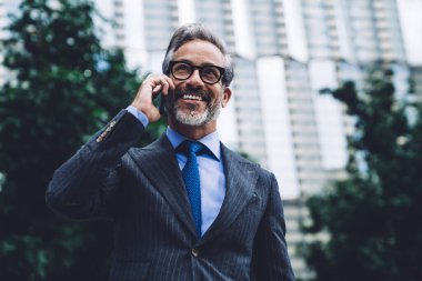 Resmi takım elbiseli ve gözlüklü neşeli, orta yaşlı, gri saçlı yakışıklı bir erkek modern dijital akıllı telefonla konuşuyor, gülümsüyor ve şehir geçmişine bakıyor.