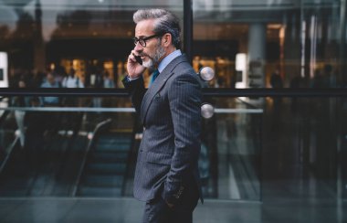 Orta yaşlı, şık, koyu renk takım elbiseli bir iş adamının cam duvarın yanında telefonla konuşurken New York 'ta elini cebine atarken yan görüntüsü.