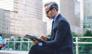 Gözlüklü ve siyah kostümlü yakışıklı iş adamının New York 'taki bulanık şehir geçmişinde tablet kullanmasının yan görüntüsü.