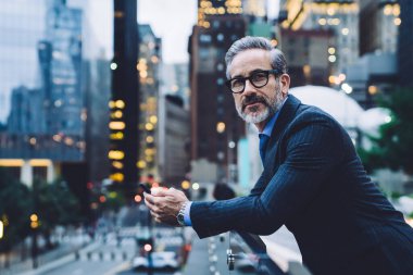 Gözlüklü ve takım elbiseli yakışıklı orta yaşlı bir adam elinde telefonla New York caddesinin arka planına yaslanıyor.