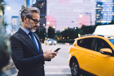 Gözlüklü ve takım elbiseli yetişkin bir erkeğin akıllı telefonuyla yazı yazıp New York yoluna karşı duruşu.