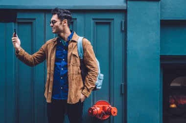 Sırt çantası ve güneş gözlüğü takmış yakışıklı genç adam New York caddesinin mavi binasında dururken gülümsüyor ve selfie çekiyor.