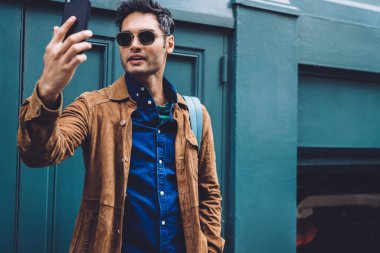 Sırt çantası olan yakışıklı, modaya uygun kıyafetler ve güneş gözlükleriyle ağzını açıp New York caddesinin mavi binasına karşı durarak selfie çeken yakışıklı bir genç.