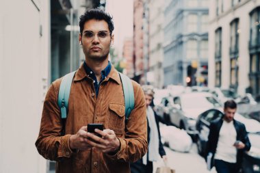 Ciddi, tıraşsız, kahverengi süet ceket giyen, kulaklık takan, New York 'ta caddede duran, akıllı telefonlu ve kameraya dikkatle bakan bir Latin.