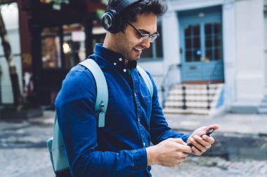Neşeli, güler yüzlü, kulaklıklı, gözlüklü, gözlüklü, akıllı telefonlu ve şehir caddesinde gündüz vakti yürürken gülümseyen yetişkin bir adamın yan görüntüsü.