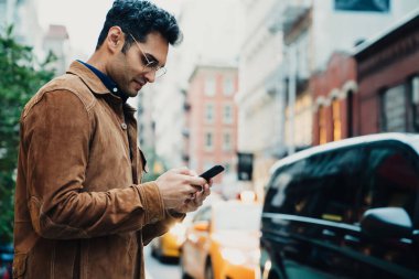 Yakışıklı, kendine güvenen, ceketli, gözlüklü, akıllı telefondan mesaj atan ve şehir caddesinde gündüz vakti ayakta duran yetişkin bir adamın yan görüntüsü.