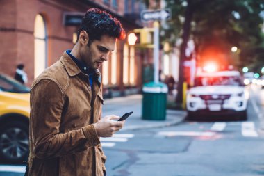 Yakışıklılığa odaklanmış, ceketli, akıllı telefon kullanan, gün ışığında trafik şeridi boyunca yürüyen esmer bir adamın yan görüntüsü.