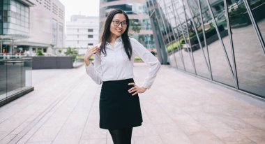 Resmi giyinmiş, gözlüklü, iş merkezinin önünde kameraya bakarak poz veren neşeli, çekici bir Asyalı kadın işçi.