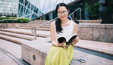 Güzel Asyalı kadın gözlüklü, büyüleyici bir şekilde gülümseyen ve ilginç kitaplar okuyan modern binanın dışındaki sınırda dinlenen.