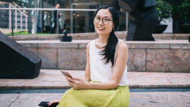 Modern kasaba caddesindeki modern binanın dışında akıllı telefon kullanırken kameraya bakıp gülümseyen gözlüklü etnik bir kadın olması güzel.
