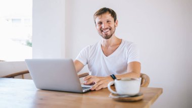 Orta yaşlı, tıraşsız, beyaz tişörtlü, beyaz duvarın yanındaki ahşap masada oturan, kahve içen ve bilgisayarda yazan basit bir erkek.