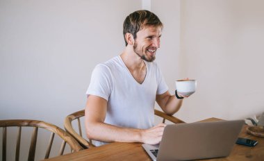 Keyifli, neşeli, sakallı bir adam. Sıcak kahve içer, evde dizüstü bilgisayar kullanır ve gözlerini başka yöne çevirir.