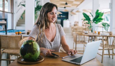 Genç milenyum kadını hindistan cevizi kokteyli ile masada oturan dizüstü bilgisayarın ayarlarını kontrol ediyor ve internet iletişimi için kafeterya web bağlantısı kullanıyor, dijital göçebe yaşam tarzı kavramı