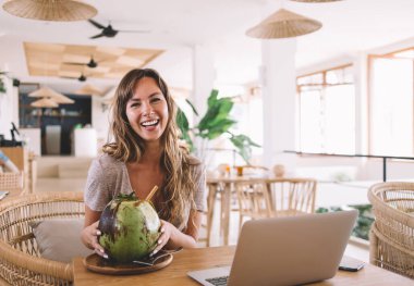 Bambu kafeteryasında egzotik tropikal içecekler içtiği için gülen neşeli Kafkasyalı serbest yazar portresi Endonezya hasır kafesinde kameraya gülümseyen mutlu kadın metin yazarı.