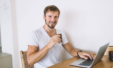 Beyaz gömlekli neşeli, sakallı, serbest bir erkek. Elinde dizüstü bilgisayarla tahta masada oturuyor. Beyaz arka planda kameraya bakıyor.