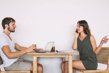 Sıradan giyinen, yakışıklı ve sakallı bir erkeğin dizüstü bilgisayarda dolaşırken beyaz arka planda akıllı telefondan konuşan neşeli, güzel bir kadının yan görüntüsü.