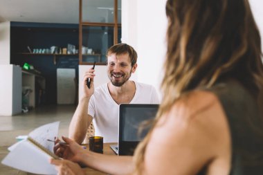 Neşeli yetişkin bir adam gülerken ve telefonla konuşurken genç bir kadın defterde yazılar gösteriyordu.