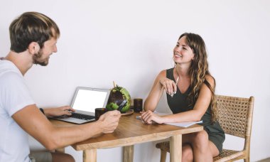 Yan bakışta, kısa elbiseli, kısa elbiseli, çapraz bacaklı, beyaz gömlekli, akıllı telefonlu kocasıyla oturan ve evinde yeşil hindistan cevizi olan ahşap masadaki boş ekranlı dizüstü bilgisayarı kullanan güzel bir kadın görülüyor.
