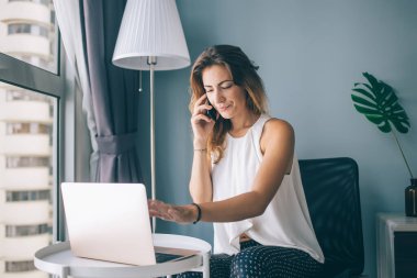 Gündelik giysiler içinde güzel bir esmer sandalyede otururken ve dizüstü bilgisayarda yazarken akıllı telefonuyla konuşuyor.