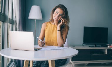 Günlük giysiler içinde gülümseyen genç esmer. Evrakları ve laptopuyla masada oturuyor. Evde kameraya bakıyor ve telefonla konuşuyor.