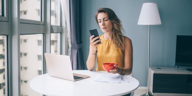 Endişeli ve endişeli kadın serbest çalışan akıllı telefon kullanırken yuvarlak masada oturuyor ve turuncu bardağı iyi aydınlatılmış bir odada tutuyor.