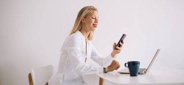 Masasında dizüstü bilgisayarı ve fincanı olan ve beyaz arka planda akıllı telefondan mesajlaşırken gülümseyen sarışın genç bayan çalışanın yan görüntüsü.