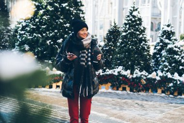 Noel ağaçlarının yanında duran ve karlı caddeye bakan, kağıt bardak ve telefonla şık kış kıyafetleri içindeki alımlı genç bayan.